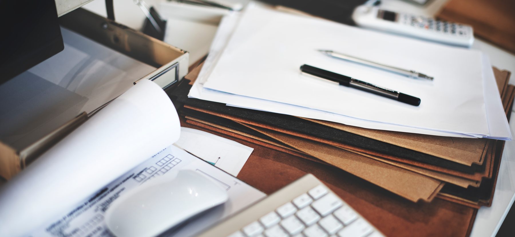 A working desk: a stack of papers with a pen resting on top, kraft folders, and the corner of a laptop