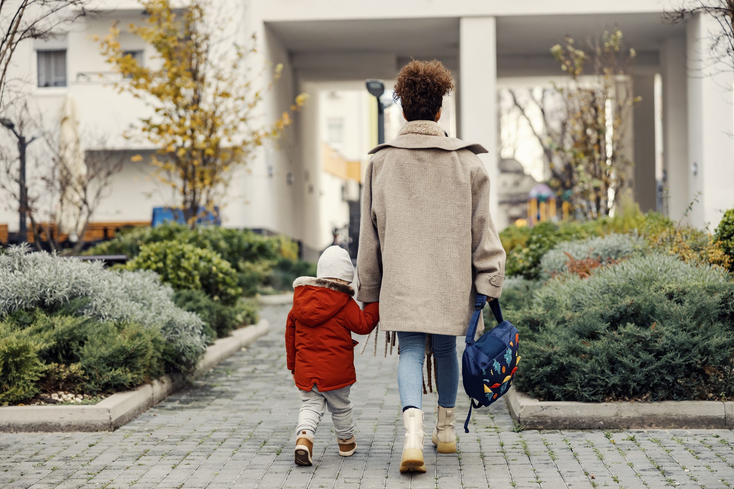 An adult and a child in a red coat walking a path between hedges, seen from behind
