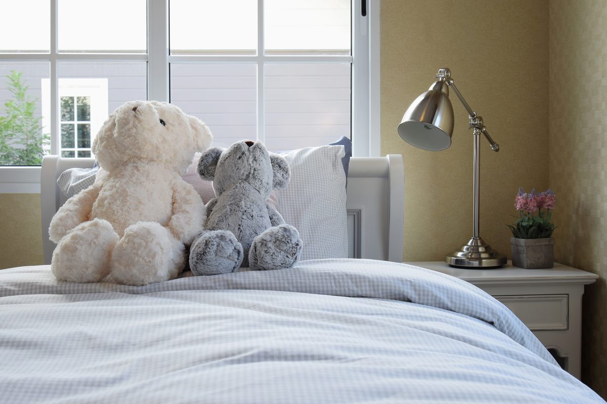 A child’s bedroom: two teddy bears against the pillows of a neatly made bed, with a bedside lamp and a small plant