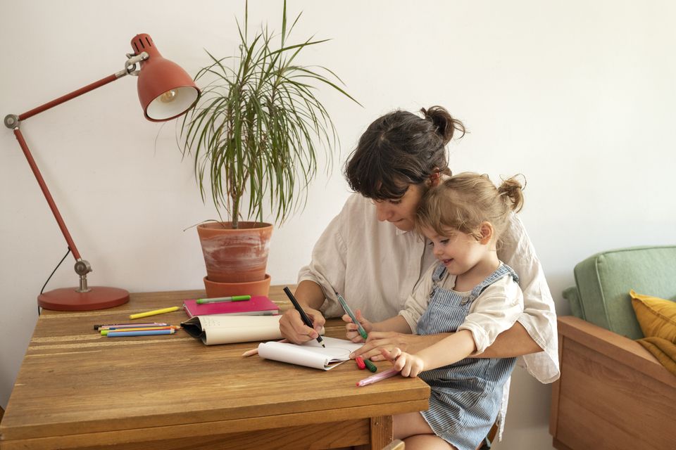 A mother and her young daughter drawing together at a desk by the window
