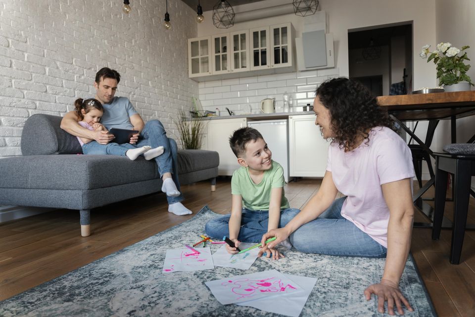 A family at home: a father and child on the sofa, a mother and son playing on the rug